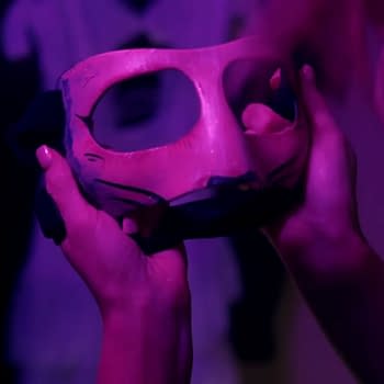 Harley Cameron holds a haunting mask with visible eye holes, surrounded by dim purple lighting, suggesting a mysterious atmosphere.