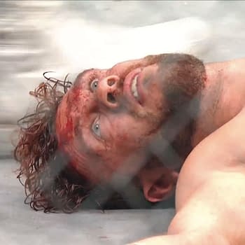 Will Ospreay, a wrestler, lies on the mat in a state of distress after a brutal Lights Out Steel Cage Match, his face covered in blood and showing visible fatigue. The scene is set against the backdrop of a steel cage.