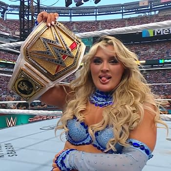 A female wrestler, Tiffany Stratton, holds up the WWE Championship belt triumphantly at WWE Summerslam. She has long blonde hair and is wearing a sparkling blue outfit.