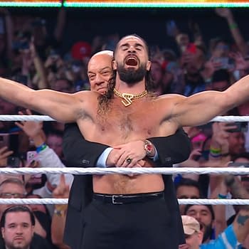 Seth Rollins celebrates in the wrestling ring, holding a championship belt high while manager Paul Heyman hugs him from behind. The crowd is cheering in the background.
