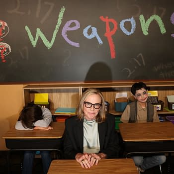 In a classroom setting, a woman with glasses sits at a desk with a serious expression, while two students exhibit contrasting behaviors: one slumped over, hidden beneath their arms, and another smiling with face paint. The chalkboard behind them displays the word 'Weapons' in colorful letters.