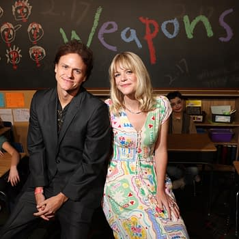 A promotional illustration featuring two actors, one wearing a black suit and the other in a colorful patterned dress, seated in a classroom setting. Behind them is a chalkboard with the word 'Weapons' written in colorful letters, with students visible in the background appearing to rest their heads on their desks.