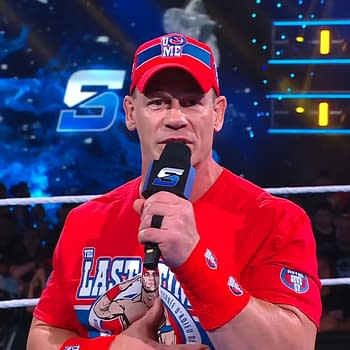 John Cena delivers a promo while holding a microphone, wearing a bright red shirt and matching cap with the words 'U Can't See Me'. The WWE SmackDown logo is visible in the background, with an energetic crowd looking on.