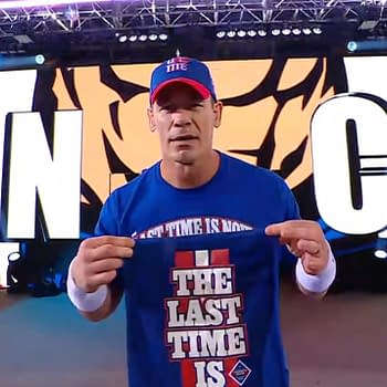 John Cena walks to the wrestling ring wearing a blue shirt that says "THE LAST TIME IS" while displaying a small item in his hand. The background features large graphics of his name.