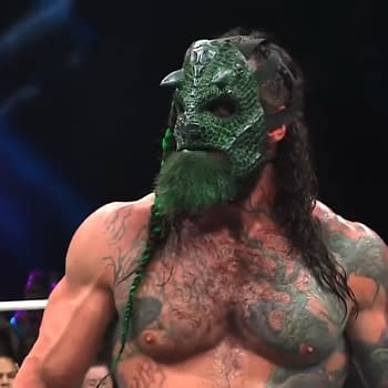 A muscular wrestler wearing a green reptilian mask with spikes and a braided hair. His upper body is heavily tattooed, and he has a green beard, standing in an arena with fans in the background.