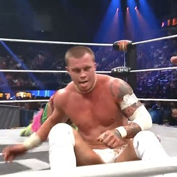 Kyle Fletcher, in wrestling gear and a determined expression, takes a moment to gloat during his match against Hiromu Takahashi. The wrestling arena is filled with lights and fans in the background.