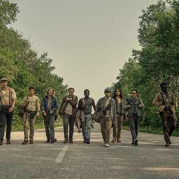 A group of nine young men walks together along a deserted road surrounded by lush greenery, each carrying bags and cameras, likely depicting a scene from the film 'The Long Walk.' The characters feature diverse clothing styles and expressions, highlighting a sense of camaraderie.