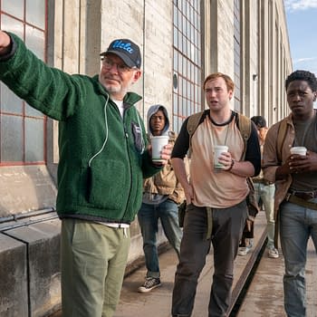 Director Francis Lawrence engages with actors Cooper Hoffman and David Jonsson on the set of 'The Long Walk', with other cast members in the background. All are holding cups, standing in front of a building with large windows.