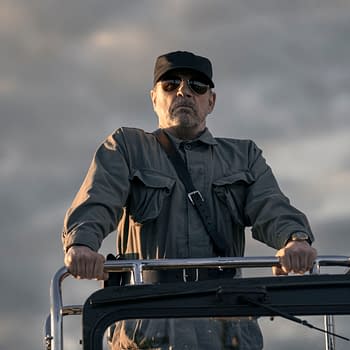 A man wearing sunglasses and a cap stands at the helm of a vehicle, dressed in a military-style shirt with pockets. He appears serious and commanding against a cloudy sky backdrop, portraying the character The Major in 'The Long Walk'.
