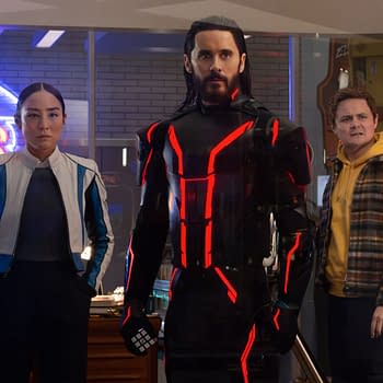 In a scene from Disney's live-action 'TRON: ARES', Greta Lee portrays Eve Kim, standing beside Jared Leto as Ares in a striking black suit with red accents, while Arturo Castro as Seth Flores looks on, all set against a vibrant arcade backdrop.