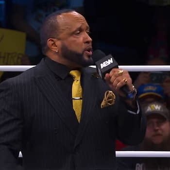 A man in a stylish black pinstripe suit with a yellow tie, MVP, passionately speaks into a microphone while standing in a wrestling ring, with fans visible in the background.