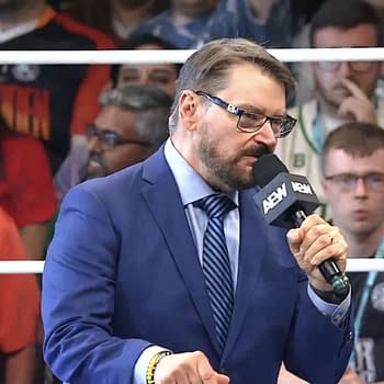 Tony Schiavone, wearing a blue suit and glasses, announces the record-breaking attendance at the AEW Forbidden Door event in London, speaking into a microphone while a diverse crowd is visible in the background.