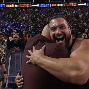Wardlow embraces Don Callis with joy, while Kazuchika Okada observes in the background at the Forbidden Door event. The audience can be seen enthusiastically reacting to the moment.