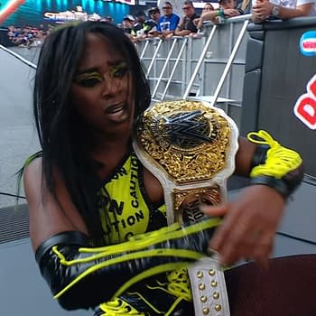 A wrestler with long hair, Naomi, is sitting on the ground after a match at WWE SummerSlam, holding a championship belt. She wears a bright yellow and black outfit with striking makeup.