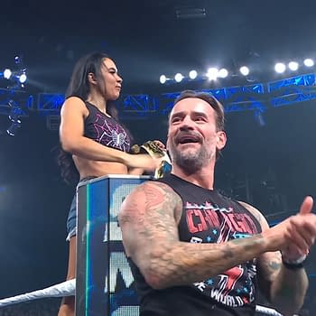 AJ Lee stands on an elevated platform, holding a championship belt and looking at the cheering crowd, while CM Punk sits beside her, clapping and smiling in response to the audience's enthusiastic reaction on WWE SmackDown.