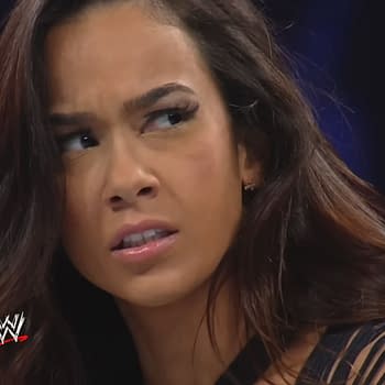 A woman with long hair, showing a concerned expression, appears in a wrestling setting. She has a serious look as she engages with the event happening around her.