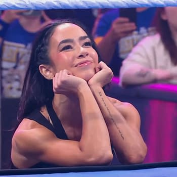 AJ Lee, a professional wrestler, sits at the edge of the ring with her chin resting on her hands, smiling warmly at the crowd during a WWE SmackDown event, with fans visible in the background. The atmosphere is vibrant and excited.