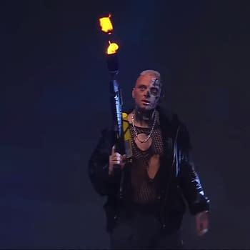 A wrestler with a heavily tattooed face and a dramatic outfit, holding a flamethrower that emits flames, appears on stage in low light, surrounded by a blue backdrop.