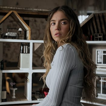 A young woman with long, wavy hair is sitting beside a detailed dollhouse, looking thoughtfully to the side. She appears to be wearing a fitted white top, enhancing a sense of contemplative emotion.