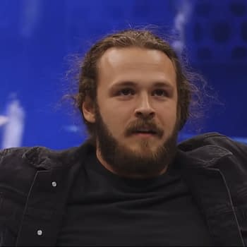 A young man with a beard and long hair sits casually, wearing a black jacket over a black shirt. The background is bright blue, hinting at a wrestling event atmosphere.
