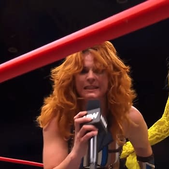 Jamie Hayter, with curly red hair and wearing a wrestling outfit, is aggressively speaking into a microphone while standing in a wrestling ring. Queen Aminata stands beside her, wearing a long yellow lace outfit and looking on.