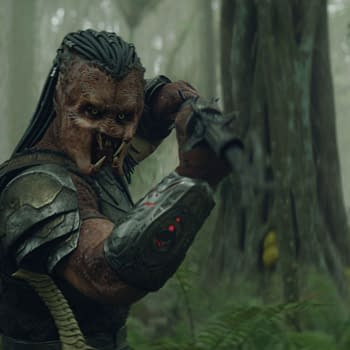 An alien character with distinct features, wearing armor and wielding a weapon, is set against a misty forest background, representing the film Predator: Badlands.