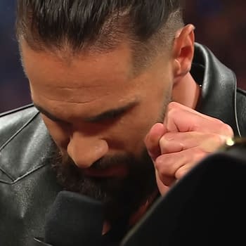 Seth Rollins is shown with his head bowed, appearing deep in thought as he prepares to deliver a scripted line during a promo on WWE Raw, wearing a black leather jacket. His hand is clenched near his face, showcasing a moment of concentration.