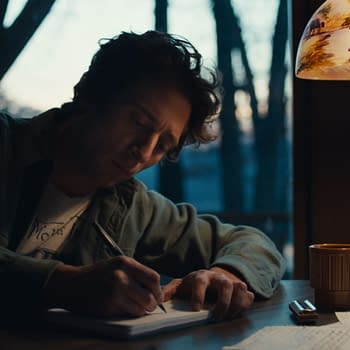 Illustration of Jeremy Allen White portraying Bruce Springsteen, seated at a table and writing in a notebook under a warmly lit lamp, with a window showing an outdoor setting in the background. This image is from 20th Century Studios' film 'SPRINGSTEEN: DELIVER ME FROM NOWHERE'.