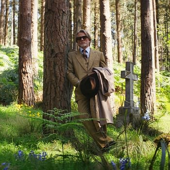 A still from 'Wake Up Dead Man: A Knives Out Mystery,' featuring a man in a tan suit and sunglasses leaning against a tree in a graveyard, surrounded by gravestones and lush greenery.