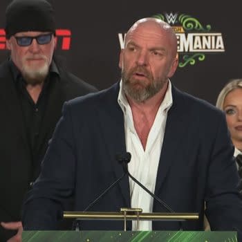 A press conference scene featuring a man in a suit speaking at a podium, with a green WrestleMania 43 logo in the background. Behind him, another man in a black outfit and sunglasses stands, along with a woman with long blonde hair dressed in a suit.