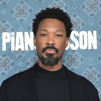 A man with curly hair and a beard is posing for the camera, wearing a black jacket and turtleneck. The background features a blue patterned design with text that reads "The Piano Lesson".