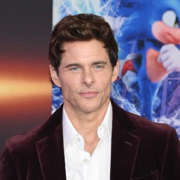A man with short, dark hair and a well-groomed appearance wears a white shirt under a dark velvet blazer. He appears at a film premiere with a colorful background featuring a blue animated character running.