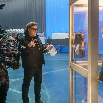 Behind-the-scenes image from 'Black Phone 2' featuring a director guiding the filming process with a camera, while a young actress stands in a glass phone booth next to an old-fashioned payphone.