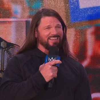 AJ Styles, wearing a black hoodie, smiles while speaking into a microphone at the Crown Jewel Perth Kickoff Show, with a championship belt displayed behind him.