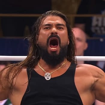 A wrestler with long hair and a beard stands in a ring, arms outstretched, passionately yelling. He wears a black tank top and a necklace, showcasing an intense expression in front of an audience.