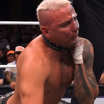 A wrestler, Darby Allin, appears to be in distress as he is choked out in the ring after a wrestling match on AEW Dynamite. The audience can be seen in the background, capturing the dramatic moment on their phones.