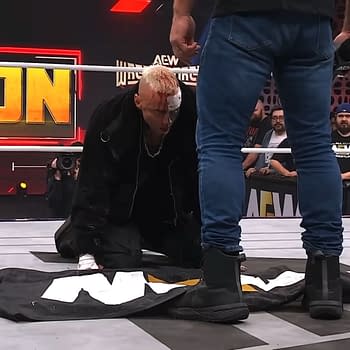 Darby Allin kneels on the wrestling ring, placing a flag on the mat as he faces Jon Moxley, who stands above him. Allin has visible injuries, adding emotional intensity to the moment.