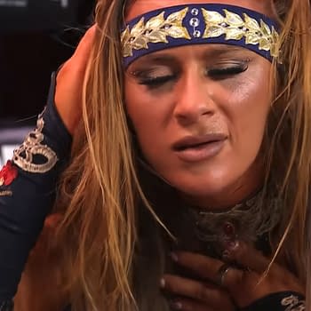 A female wrestler shows signs of distress, holding her head with a concerned expression while wearing a blue and gold headband and a decorated costume. Her makeup is slightly smudged, indicating the intensity of the moment.