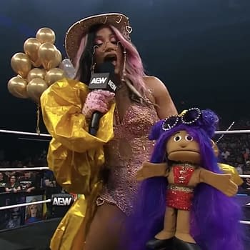 Mercedes Moné stands in the wrestling ring holding a microphone and a colorful puppet, Mini Moné, while wearing a sparkling outfit and surrounded by balloons.