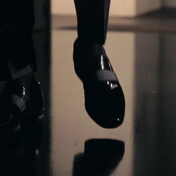 A close-up of a foot wearing a shiny black shoe, poised to take a step on a reflective surface. The shoe features a strap, emphasizing a sleek design.