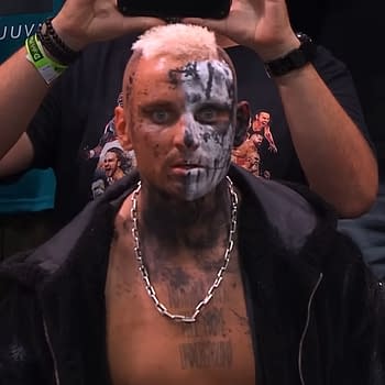 Darby Allin, a professional wrestler, shows off a striking look with half of his face painted in black and white and wearing a silver chain. He sits in the audience, visibly focused, with fans in the background.
