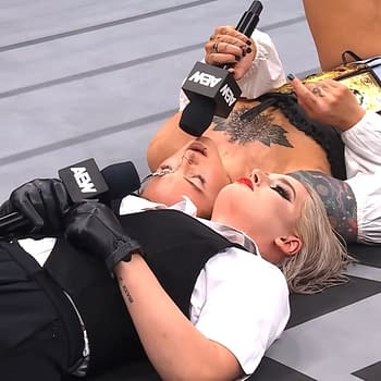 Toni Storm and Kris Statlander chat while lying on the wrestling ring floor, each holding a microphone. Statlander has a championship belt next to her.