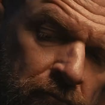 Close-up of a man with a beard and intense expression, wearing a suit. The background features a blurred figure, contributing to a dramatic cinematic moment.