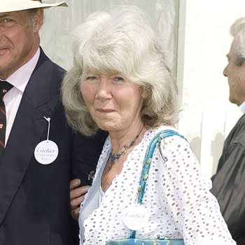 Dame Jilly Cooper, a notable author, arrives at the Cartier International Polo event in Windsor, wearing a white patterned dress and holding a blue handbag. She is accompanied by a man in a suit and hat, both sporting event badges.
