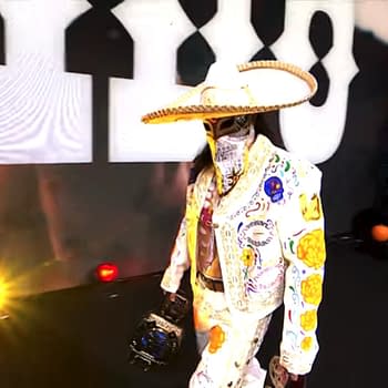 A masked wrestler wearing a colorful, intricately designed outfit and a large sombrero walks toward the ring, holding a championship belt. The backdrop features bright lights and a playful, festive atmosphere.