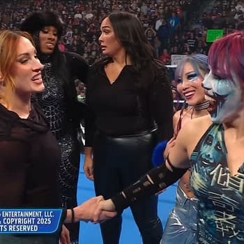 Five WWE female wrestlers are interacting in the wrestling ring, with one wrestler, Becky Lynch, shaking hands with another. The background features an enthusiastic crowd, with various fans visible.
