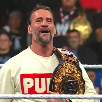 CM Punk stands in a wrestling ring wearing a white sweater with 'PUNK' printed on it and holding a championship belt, looking happy and relaxed. The audience is visible in the background, showing excitement.