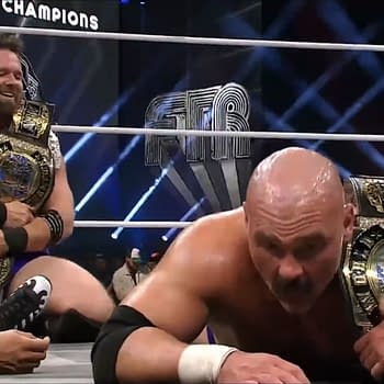 Two wrestlers are celebrating in the ring after winning the men's tag titles at AEW Full Gear. One is holding a championship belt while the other is on the mat with a belt draped over him.