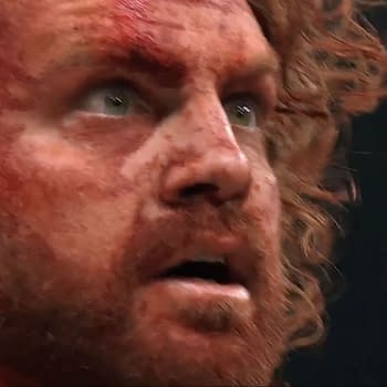 A close-up of a man with curly red hair and a beard, showing a shocked and distressed expression, covered in blood, likely reacting to a recent event in a wrestling match.