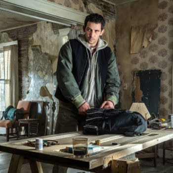 A scene from the film 'Disclosure Day,' featuring a man, Josh O'Connor, packing a black bag on a wooden table in an aged, unkempt room with floral wallpaper and a mix of furniture in the background.